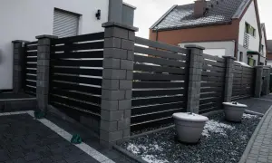 Nowoczesne ogrodzenie z czarnych paneli i kamiennych słupków. Jego wysokość zapewnia prywatność. Dwa duże donice z zimowym akcentem śniegu.