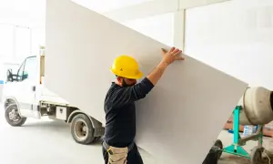 Budowlaniec w żółtym kasku niesie dużą płytę, która posłuży do budowy ścianki działowej z płyty kartonowo gipsowej.