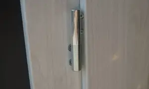 Metalowy zawias drzwi, widoczny na jasnym, drewnianym tle. Może być potrzebna regulacja zawiasów drzwi, aby zapewnić płynne otwieranie.