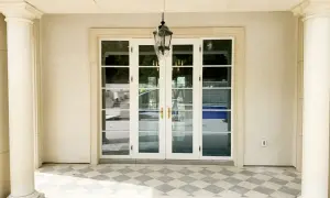 Eleganckie drzwi francuskie z białymi ramami i złotymi klamkami, prowadzące do ogrodu z basenem.