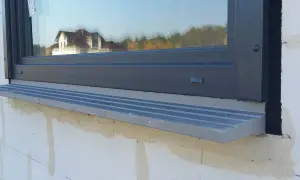 Szary parapet pod oknem, którego framuga odbija dom i drzewa.