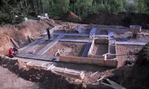 Budowa fundamentów domu. W wykopie widać zbrojenie i szalunki. Pracownicy przygotowują teren pod budowę, a chudziak czeka na beton.