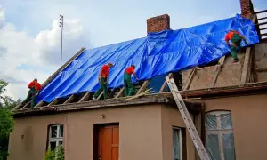 Pracownicy na dachu, przykrytym niebieską plandeką, wykonują prace budowlane. Może to być próba ukrycia **zmiany konstrukcji dachu bez pozwolenia**.