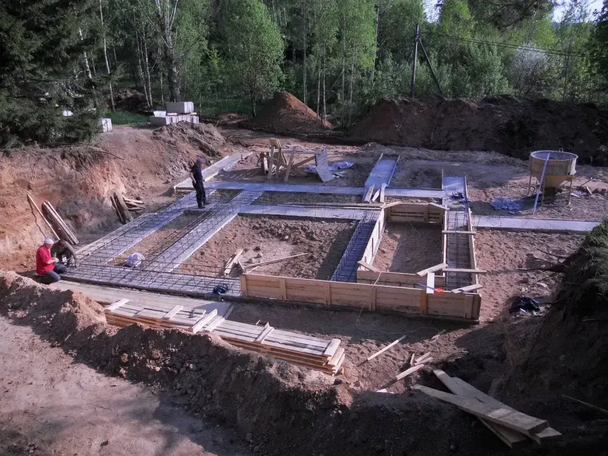 Budowa fundamentów domu. W wykopie widać zbrojenie i szalunki. Pracownicy przygotowują teren pod budowę, a chudziak czeka na beton.