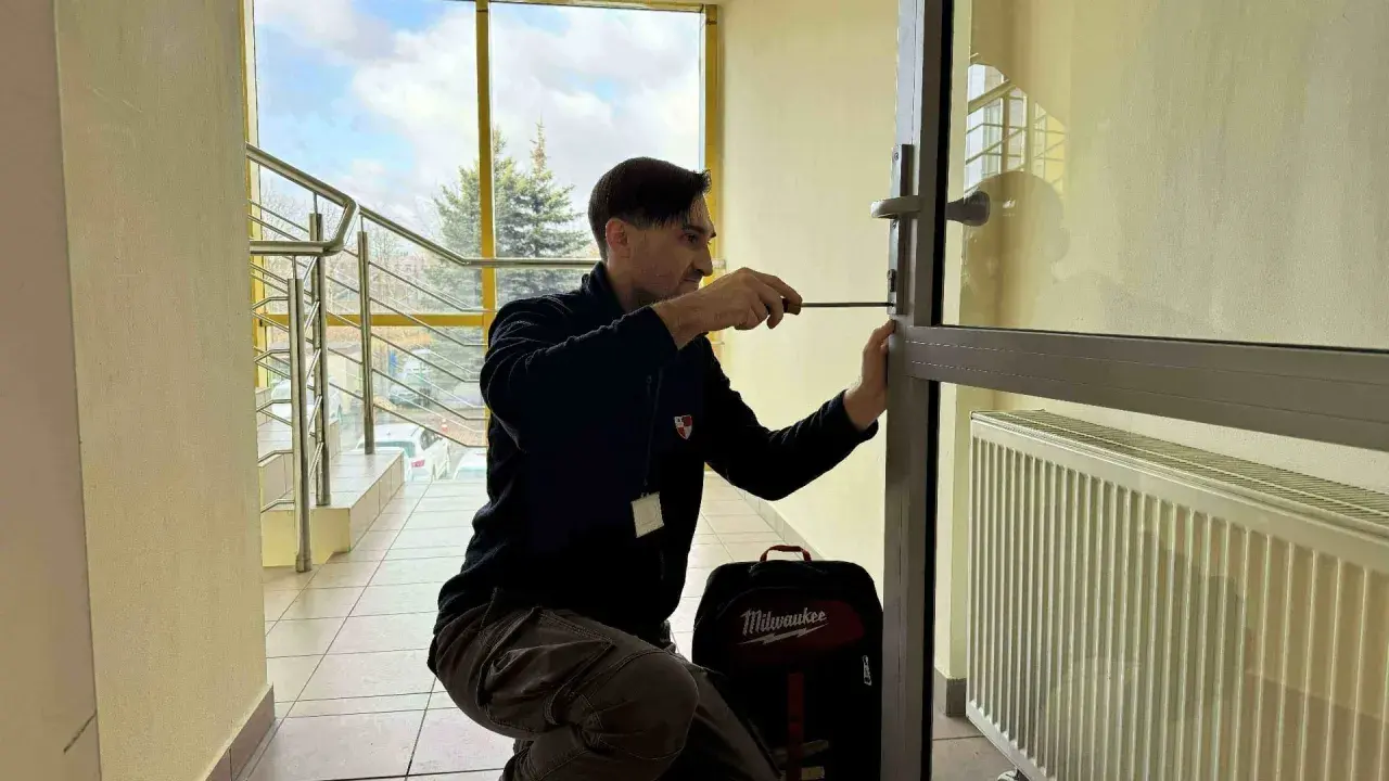 Mężczyzna próbuje otworzyć drzwi bez klucza, używając śrubokręta. Obok niego stoi torba z narzędziami.