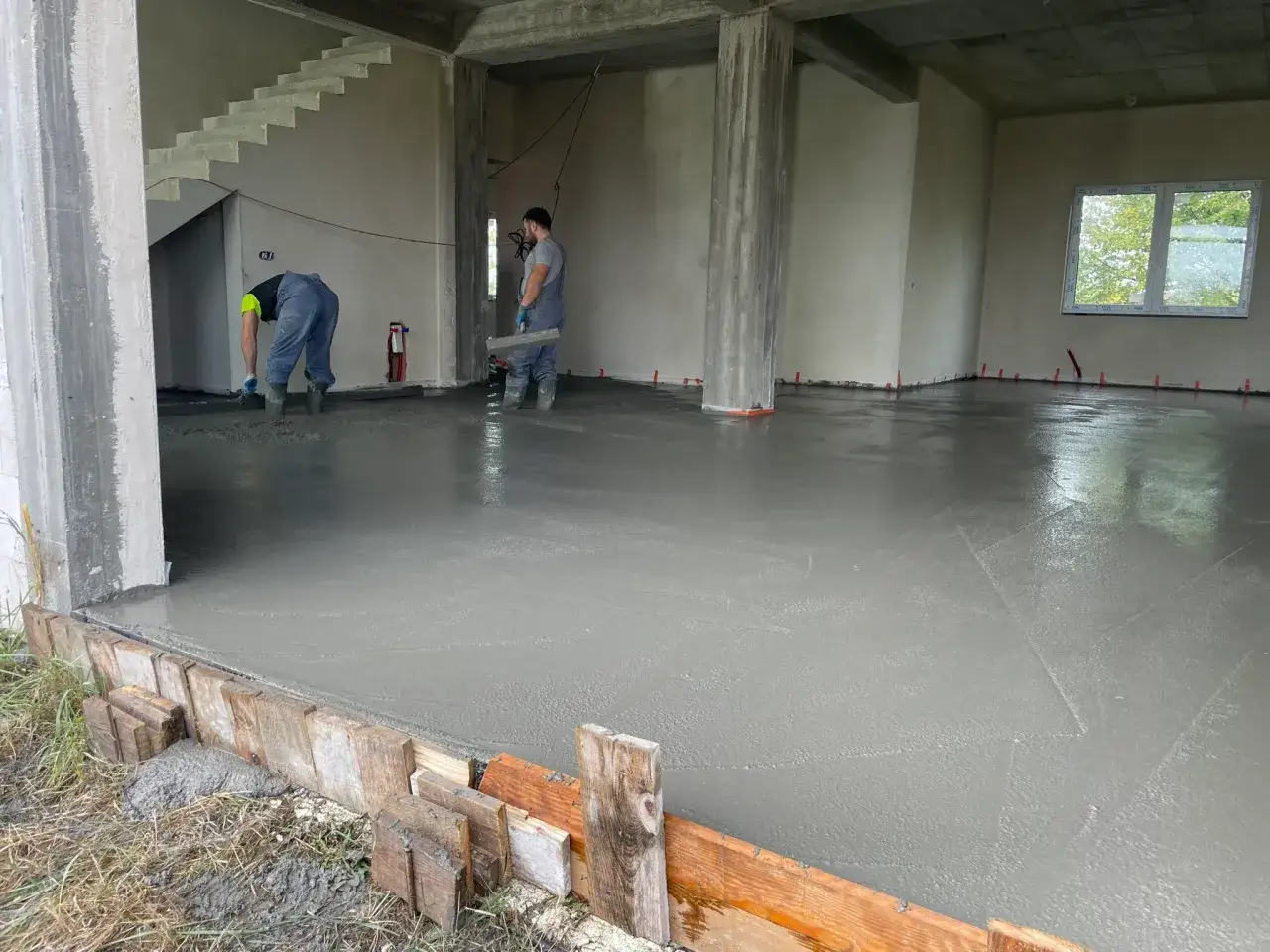 Pracownicy wykańczają świeży, gładki beton C25/30 na posadzce. W tle widać schody i okno.