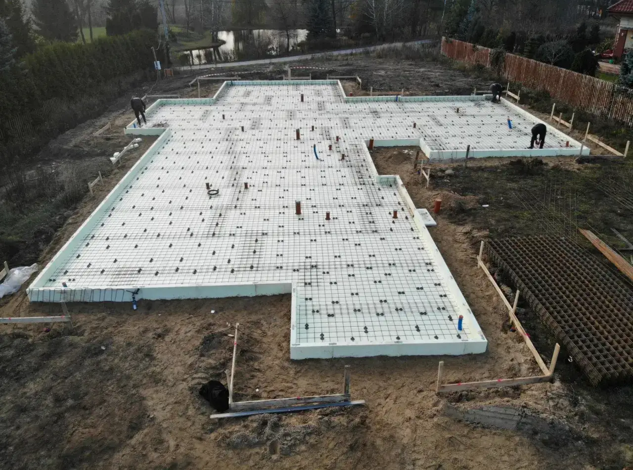 Fundament pod budowę domu z wykorzystaniem betonu C25/30. Widoczna zbrojona płyta fundamentowa z izolacją styropianową i systemem rur.