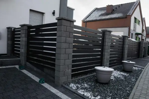 Nowoczesne ogrodzenie z czarnych paneli i kamiennych słupków. Jego wysokość zapewnia prywatność. Dwa duże donice z zimowym akcentem śniegu.