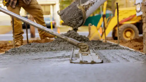 Pracownik wylewa beton na chudziaka, używając kielni do wyrównania powierzchni. W tle widać koparkę.