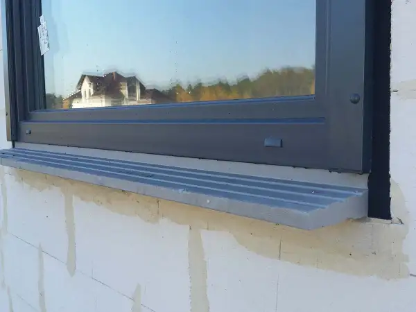 Szary parapet pod oknem, którego framuga odbija dom i drzewa.