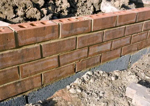 Ceglana ściana ogrodzenia z widocznym szarym bloczkiem fundamentowym, który stanowi alternatywę dla tradycyjnej podmurówki.