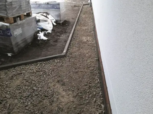 Budowa opaski wokół domu z kamienia. W tle palety z materiałami budowlanymi i resztki śniegu.